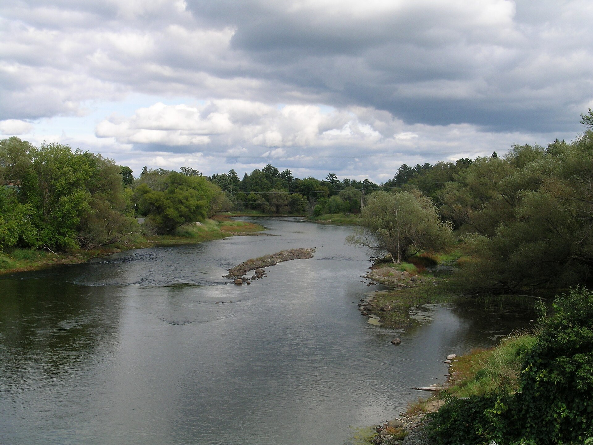 The Mississippi River flows between dense green banks near Grand Rapids, Minnesota, beneath a cloud-filled sky.